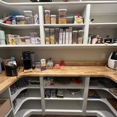 Organized kitchen pantry with clear containers, snacks, appliances, and wooden countertops in a white shelving unit.