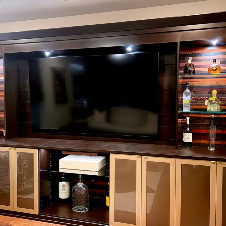 Modern entertainment center with large TV, glass shelves displaying liquor bottles, and wooden cabinetry with lights.