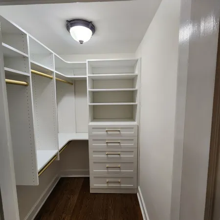 Bright walk-in closet with white shelves, drawers, gold hanging rods, and wooden floor under a ceiling light.