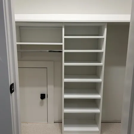 Empty white closet with multiple shelves, a small door with a black knob, and a hanging rod inside a carpeted room.