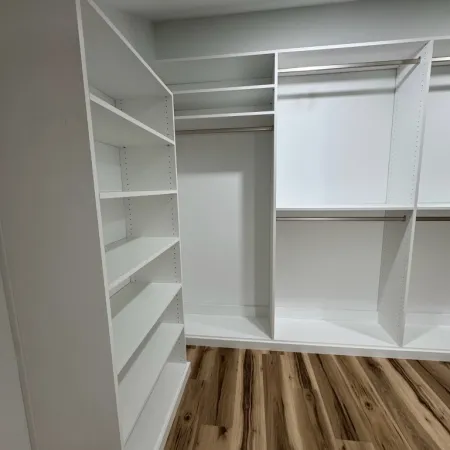 Empty white walk-in closet with shelves and hanging rods on wood style laminate flooring.