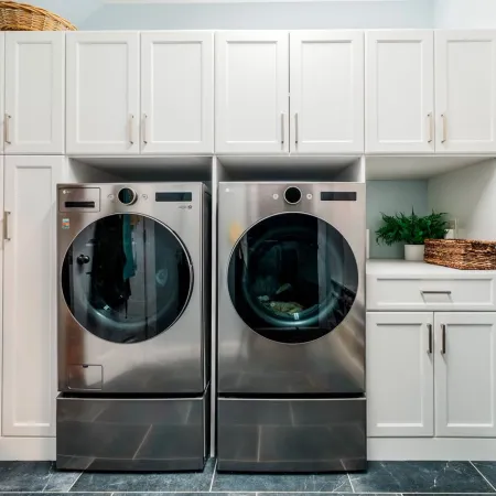 Modern laundry room with stainless steel washer and dryer, white cabinetry, wicker baskets, and gray tile floor.