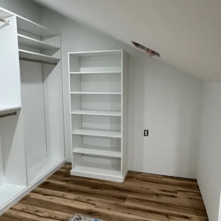 Empty white closet with built-in shelves and hanging rods on wooden floor under a sloped ceiling.