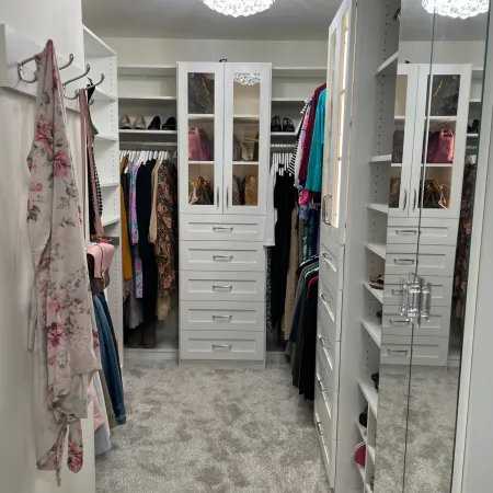 Organized walk-in closet with white shelves, drawers, handbags, hanging clothes, and soft gray carpet flooring.