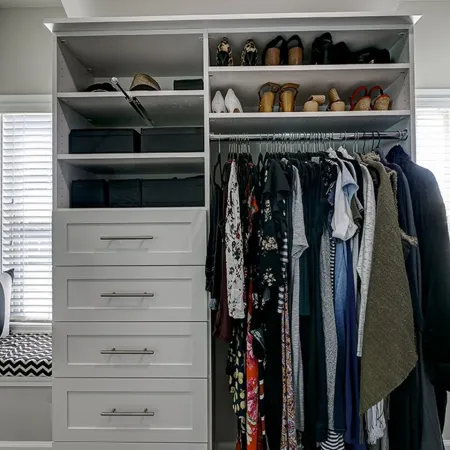 Organized white walk-in closet with hanging clothes, shoe shelves, drawers, and window seating with cushions.