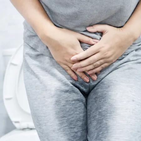 Person in gray clothes holding lower abdomen in bathroom near toilet experiencing discomfort or pain.
