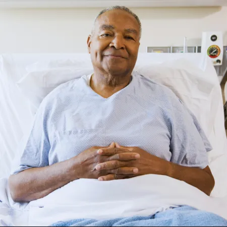 Smiling elderly man resting in hospital bed wearing light blue gown with hands folded over blanket.