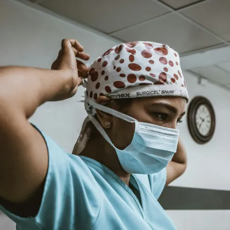 Healthcare worker wearing surgical mask and polka dot scrub cap adjusts head covering in hospital hallway.