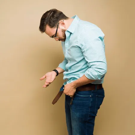 Man in blue checkered shirt and jeans adjusting his brown belt against beige background.