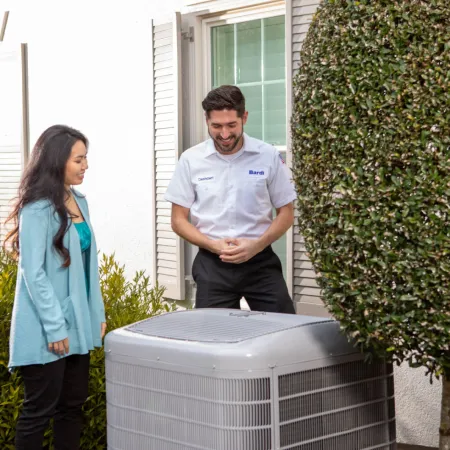 Technician explaining HVAC unit to woman outside house near trimmed tree and bushes on sunny day