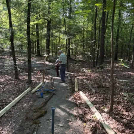 Man working on building a wooden frame pathway through a dense green forest with scattered tools.