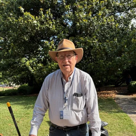 Elderly man in hat and glasses standing outdoors with tools and a workbench on a sunny day.