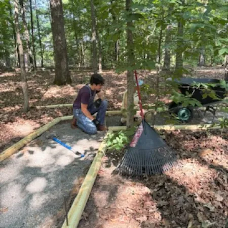 Man kneeling and working on wooden frame pathway in wooded area with rake and wheelbarrow nearby