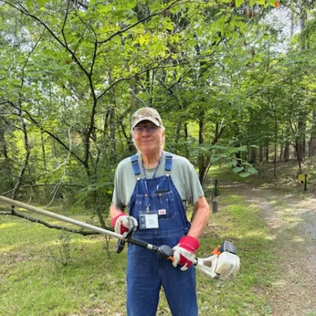 Man in overalls and gloves holding a brush cutter in a wooded outdoor area with green trees and grass.