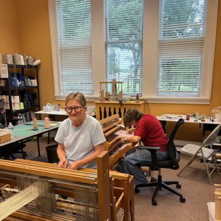 Two women working on wooden weaving looms in a bright, cozy craft room with large windows and yarn shelves.