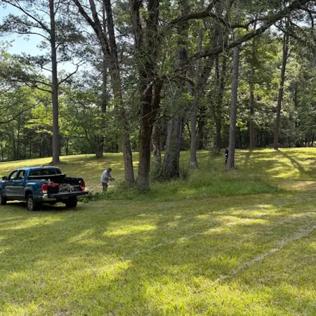 Blue pickup truck parked on grassy field near trees with person working by truck bed on sunny day