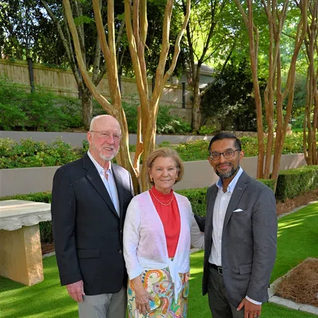 Three adults standing on green grass in a garden with trees and a stone bench, smiling at the camera.