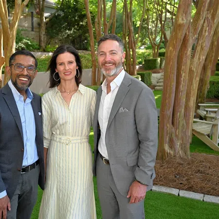 Three professionally dressed people standing outdoors in a garden with trees and greenery, smiling at the camera.