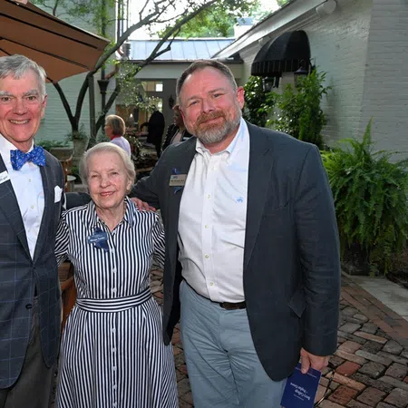 Three formally dressed people smiling outdoors at a garden event with brick flooring and patio furniture.