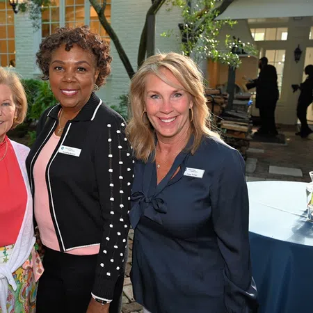 Three women smiling outdoors at a social event with name tags, standing on a brick patio near a building.