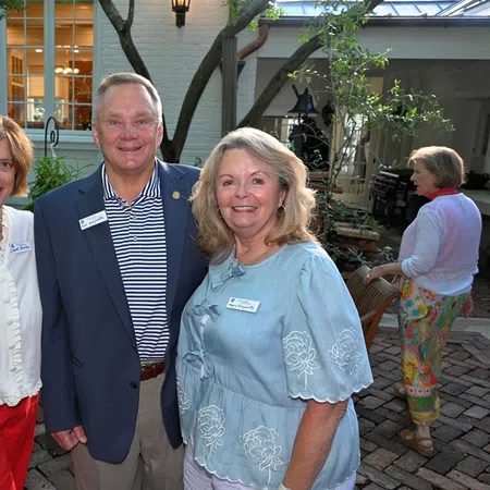 Three adults smiling outdoors on a brick patio near a house during a social gathering at dusk.