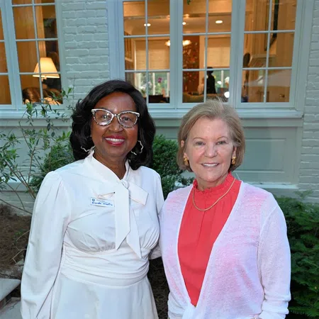 Two women smiling outdoors, one in white blouse and green polka-dot pants, the other in colorful skirt and coral top.