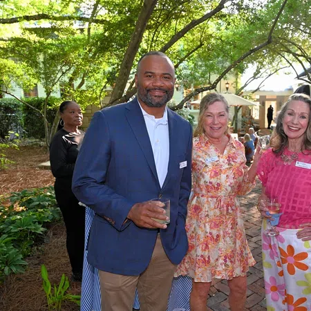 Group of adults socializing outdoors at a garden event with drinks in hand and greenery around.