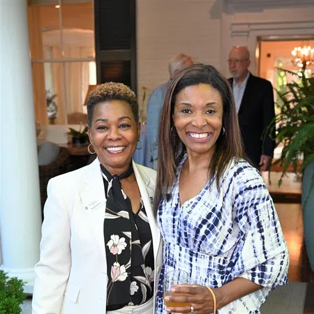 Two smiling women at a social event, one in a white suit and floral blouse, the other in a blue and white dress holding a drink.