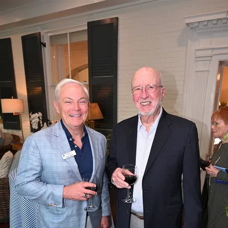 Two older men in suits smiling and holding glasses of red wine at an indoor social gathering.