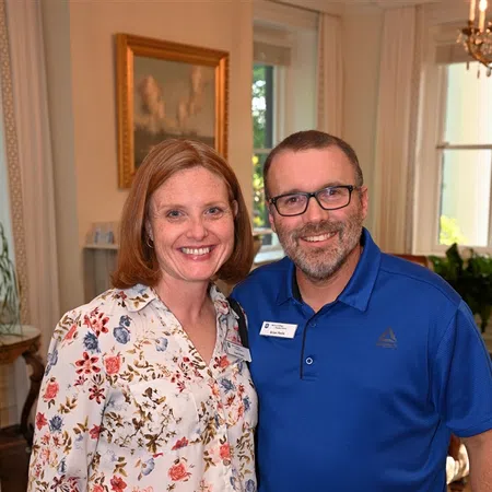 Smiling man and woman standing together indoors at a social event, woman holding a drink, both wearing nametags.