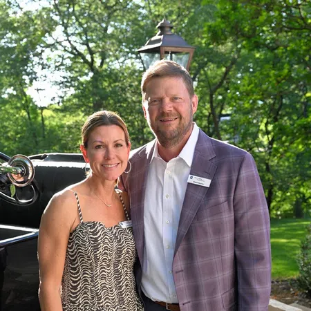 Smiling couple standing outdoors on brick path with vintage car and greenery in the background, wearing formal attire