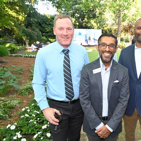 Three professionally dressed men smiling outdoors at a garden event with greenery and guests in the background