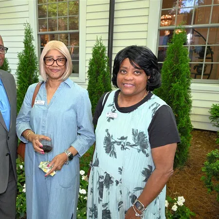 Three people posing outdoors at a social event, dressed in semi-formal attire with name tags, greenery in background