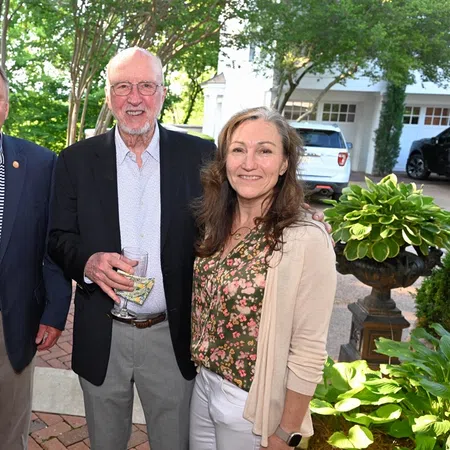 Three adults smiling outdoors near greenery and parked cars, two men in blazers and a woman in floral top and cardigan.