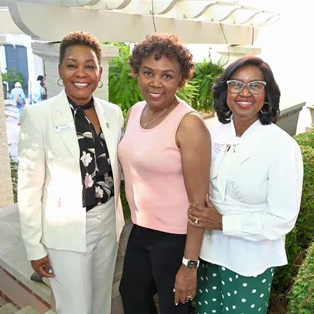 Three women smiling outdoors near greenery and brick steps, dressed in business casual and casual attire.