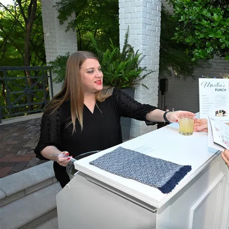 Woman serving a drink to another woman at an outdoor white bar with greenery and a Mother's Punch menu