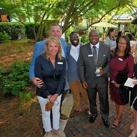 Group of diverse professionals posing and smiling outdoors at a garden social event under trees.