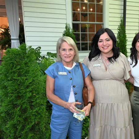 Three women smiling outdoors near greenery and a building with large windows during daytime.