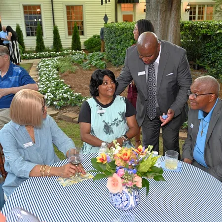 People socializing outdoors at a garden party around a striped table with a floral centerpiece in daytime.