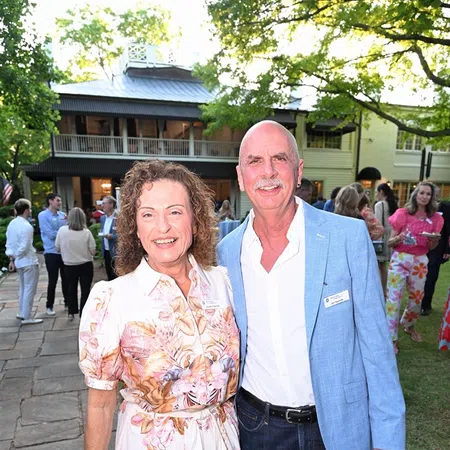 Middle-aged couple smiling at outdoor social gathering in front of large house with trees and guests around.