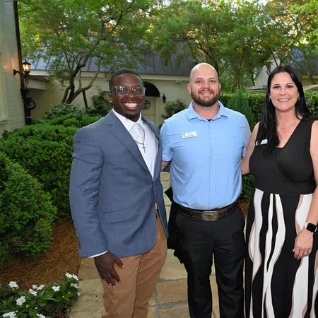 Four diverse professionals smiling outdoors in business casual attire with greenery and flowers in the background.