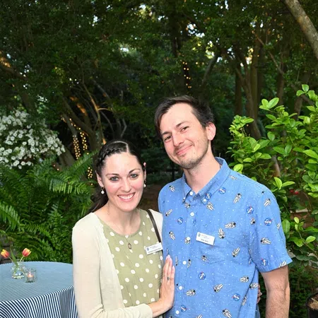 Smiling couple standing outdoors in garden setting with string lights and greenery in background.