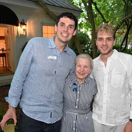 Two young men and an elderly woman smiling outdoors near a white building with trees and string lights in the background