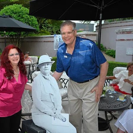 Man and woman posing outdoors with a grayscale statue of a seated person, surrounded by people and patio furniture.