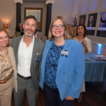 Three professionally dressed people smiling at a networking event in a well-lit room with artwork and informational posters.