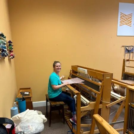 Smiling woman weaving fabric on a large wooden loom in a cozy craft room with thread spools on the wall.