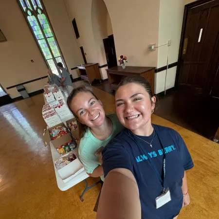 Two smiling young women take a selfie at a food donation table inside a church with stained glass windows.