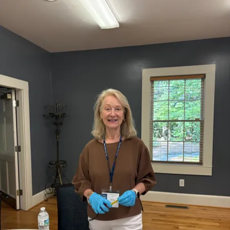 Smiling woman wearing blue gloves standing in a room with wooden floor and window, holding a name badge.