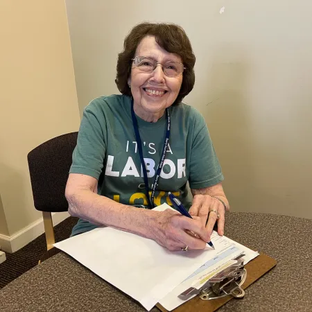 Smiling elderly woman wearing glasses and a green Labor Day shirt writing on clipboards at a round table.