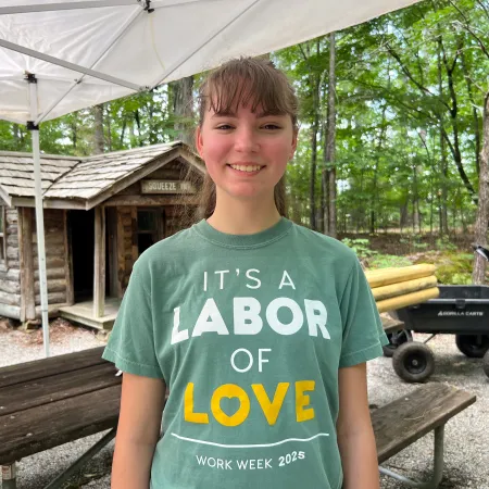 Young woman wearing a green shirt that says It’s a labor of love at an outdoor rustic work site with wooden cabins and trees.